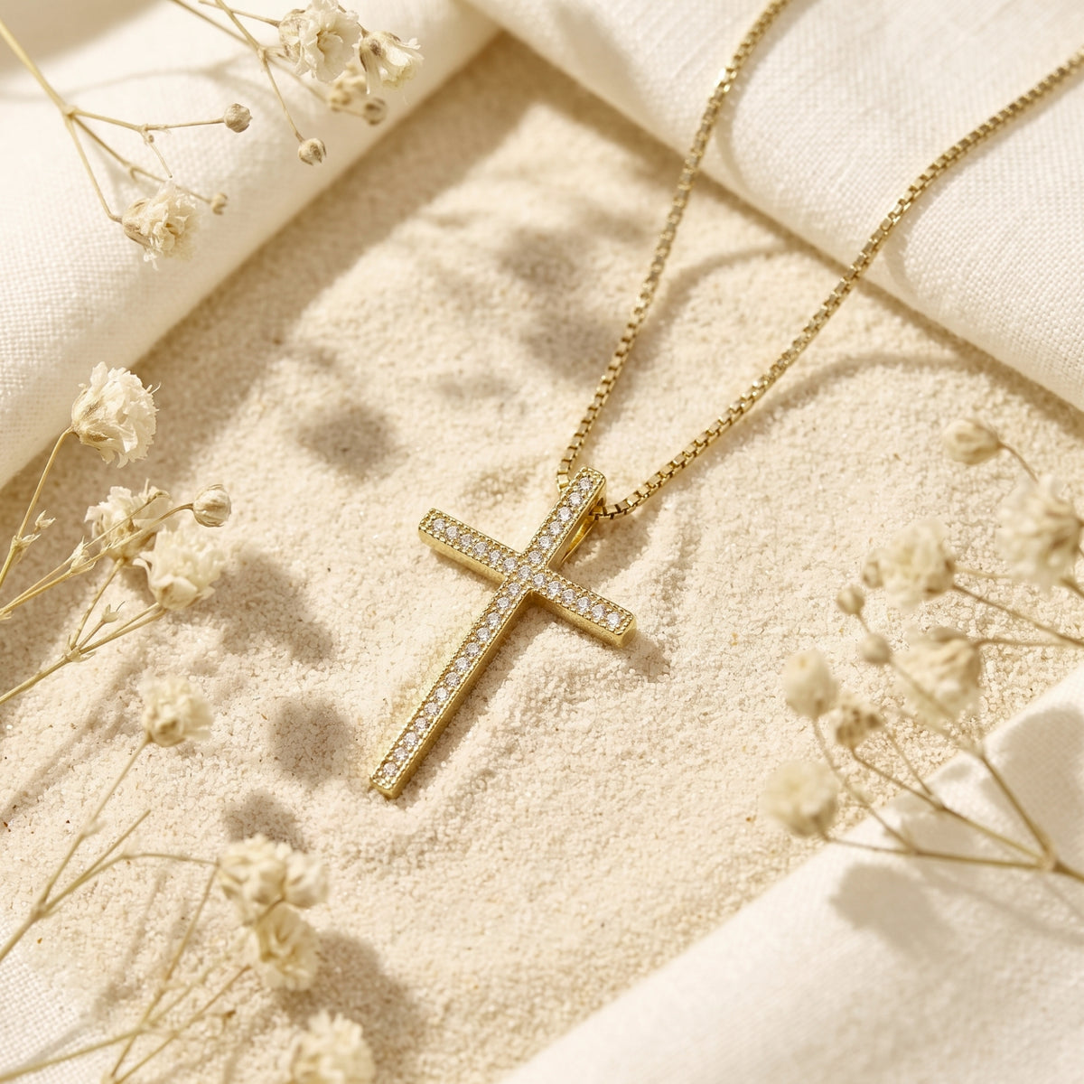 Gold-tone cross necklace with pavé stones laid on fine sand among white dried flowers.