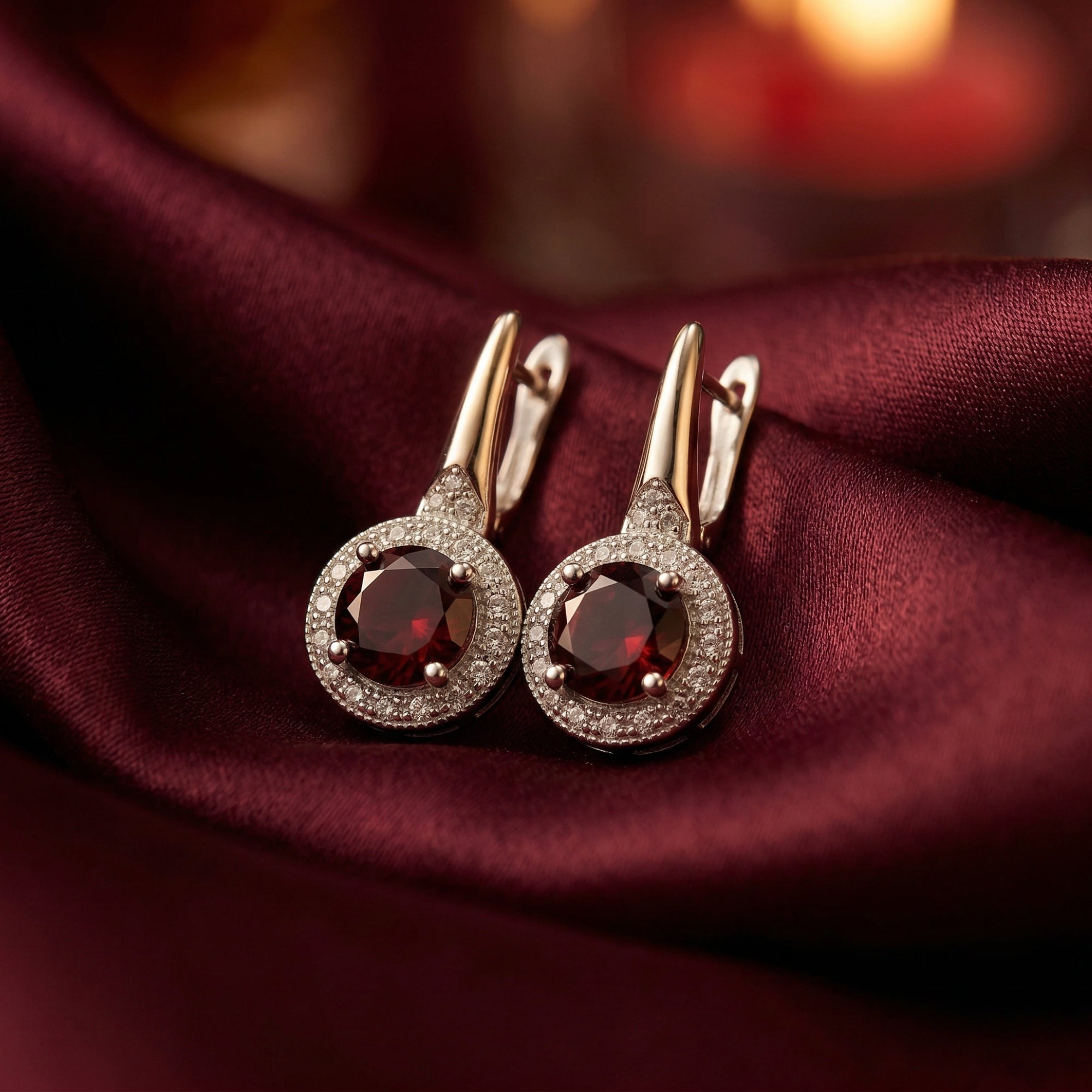 Round cut garnet drop earrings in sterling silver with a halo setting on burgundy silk fabric.