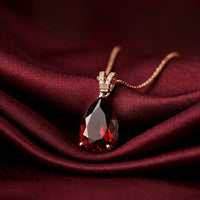 Pear cut garnet pendant in 18K rose gold on a matching chain, resting on draped burgundy silk fabric.
