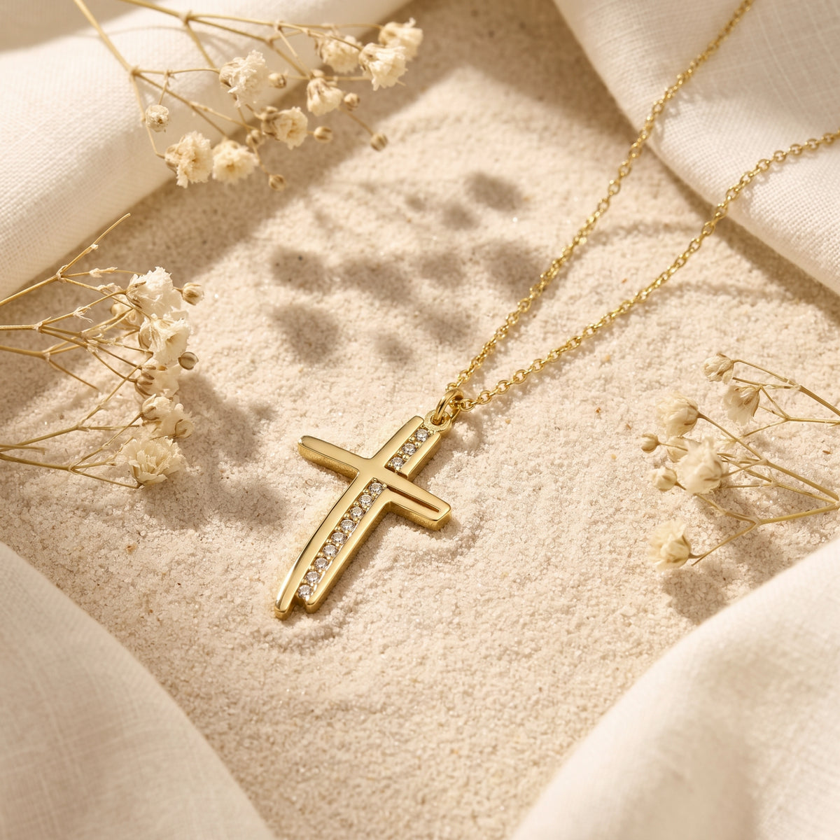 Gold-tone channel cross pendant with clear cubic zirconia accents resting on light sand with dried flowers.