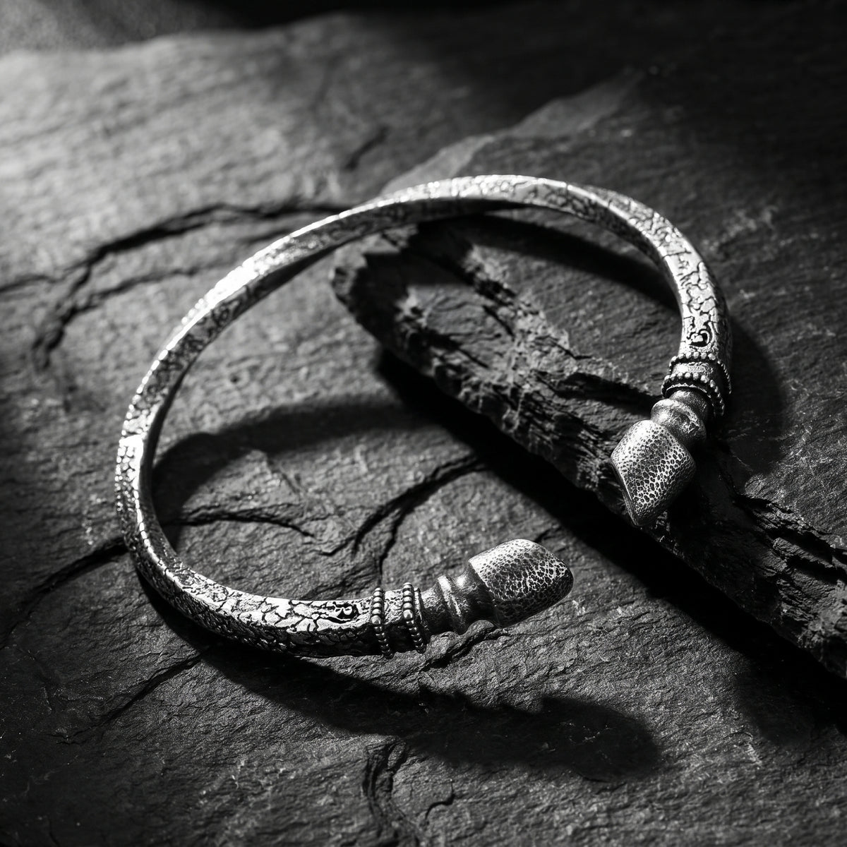 Angled close-up of an oxidized silver open cuff bracelet with textured finial ends on dark slate.