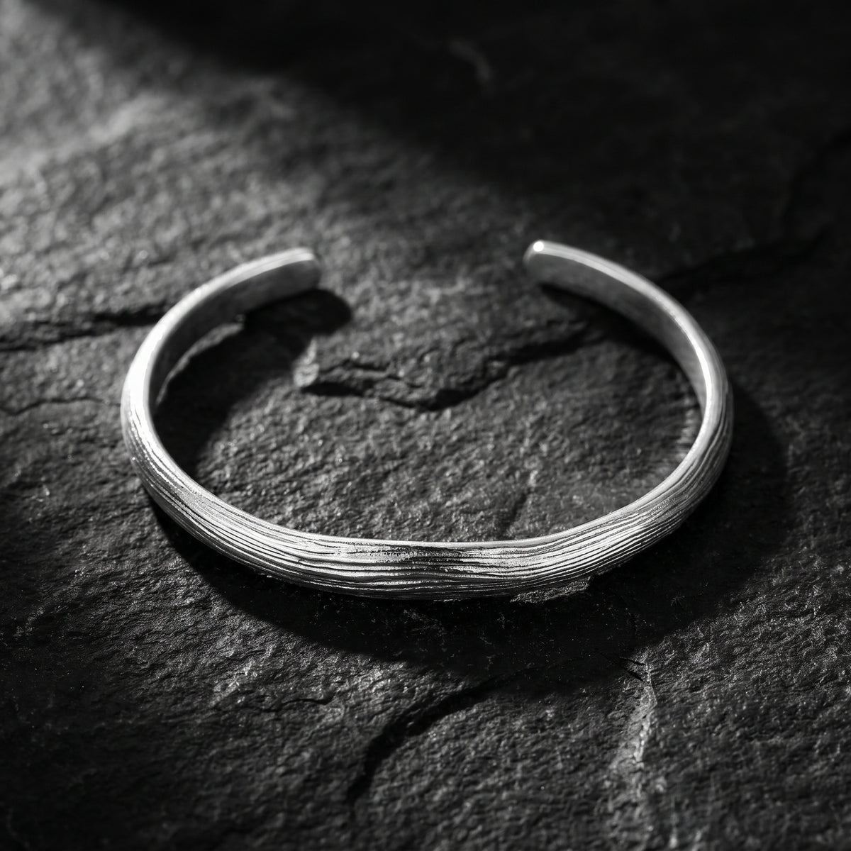 Silver open cuff bracelet with bark-textured ridges displayed on a dark slate surface.