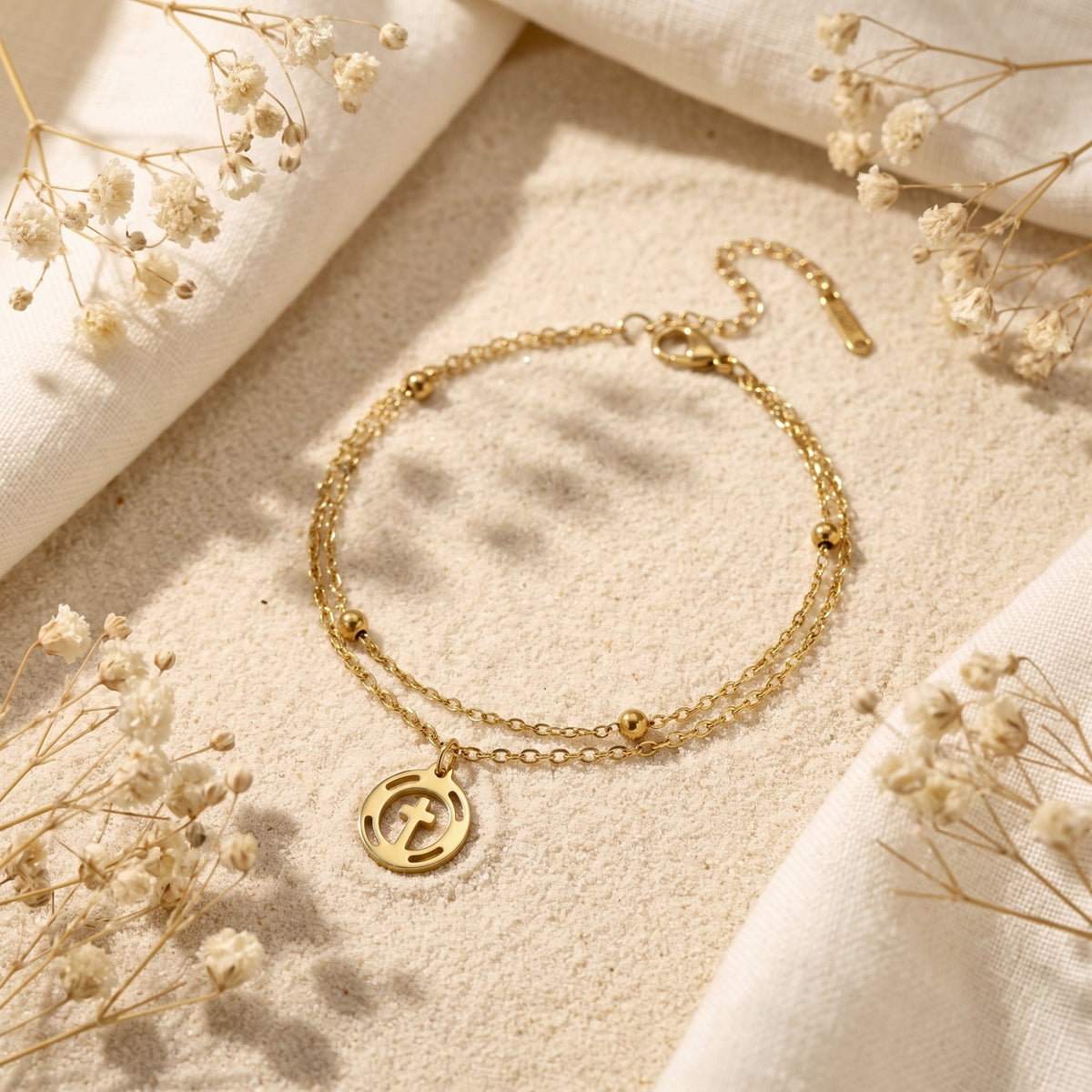 Gold-tone double-strand bracelet with a circular cross charm and beaded chain, displayed on sand with dried flowers.