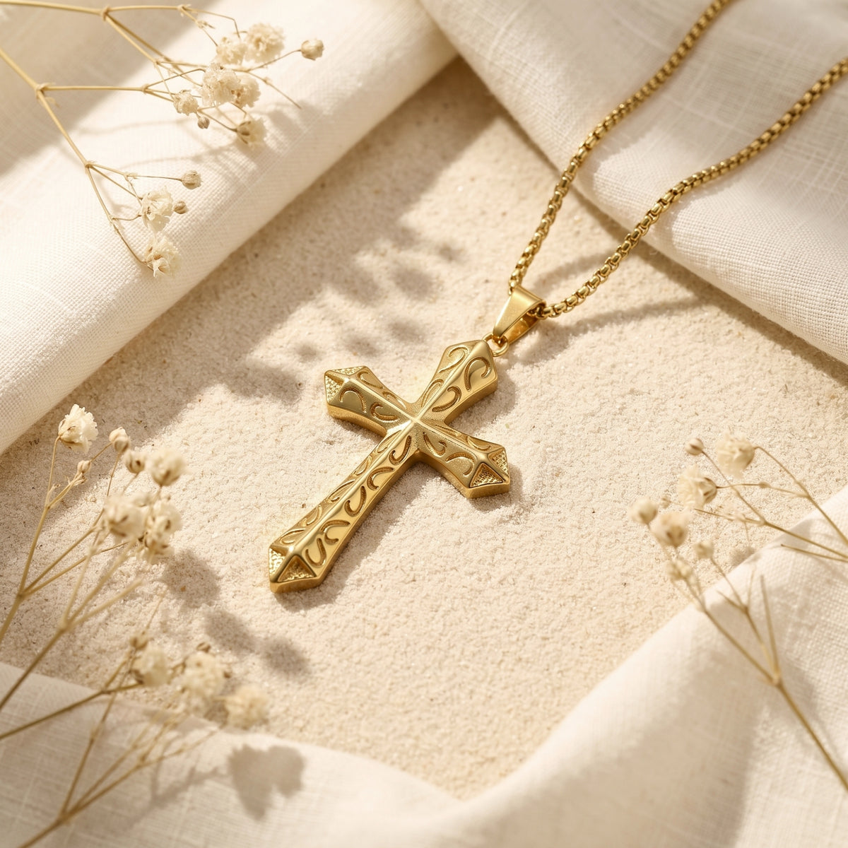 Gold-tone scrollwork cross pendant resting at an angle on sand, surrounded by white fabric and dried flowers.