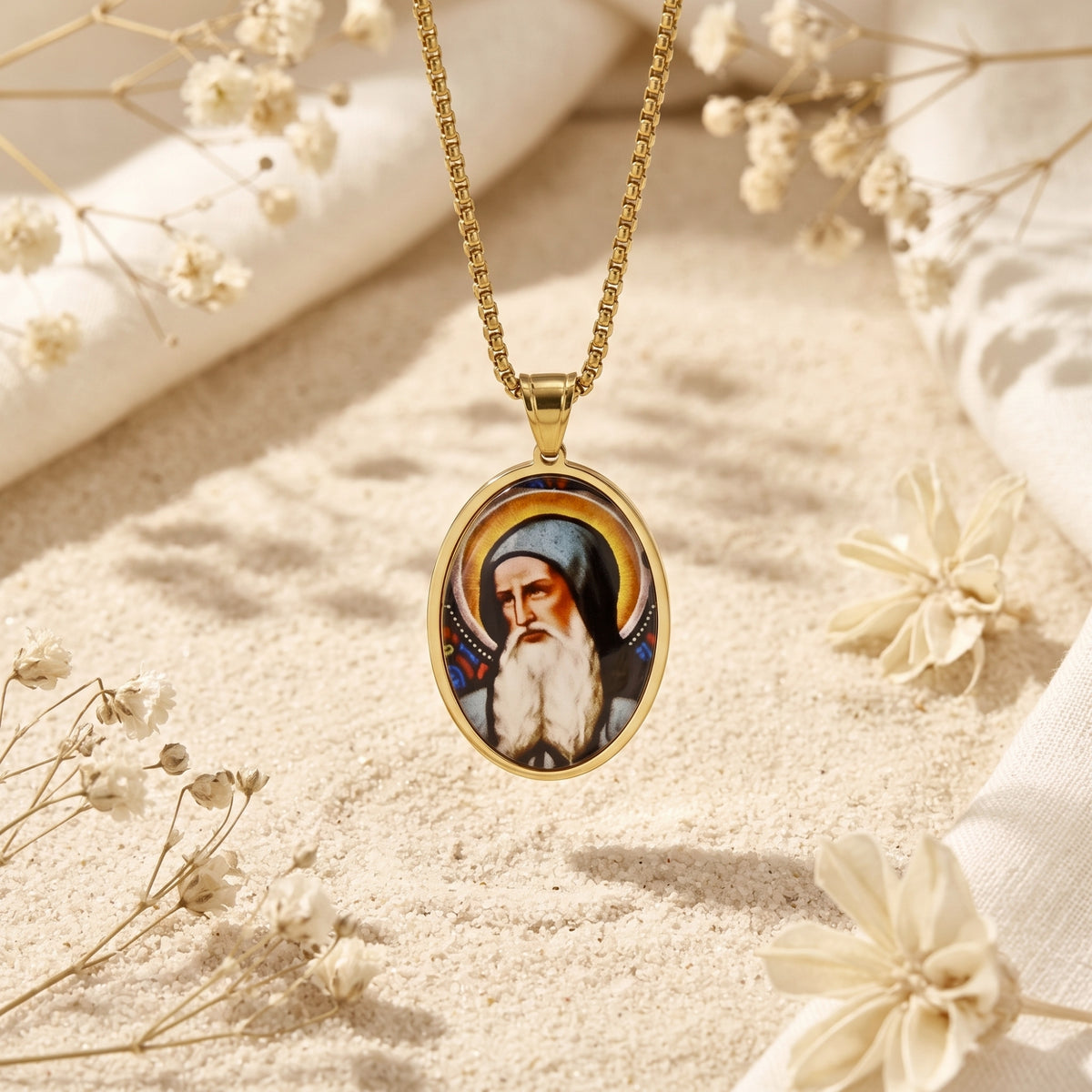 Gold-tone oval saint icon pendant hanging straight against sand with dried floral accents.