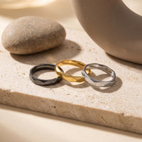 Trio of faceted stainless steel stackable rings in black, gold, and silver on a stone lifestyle background.