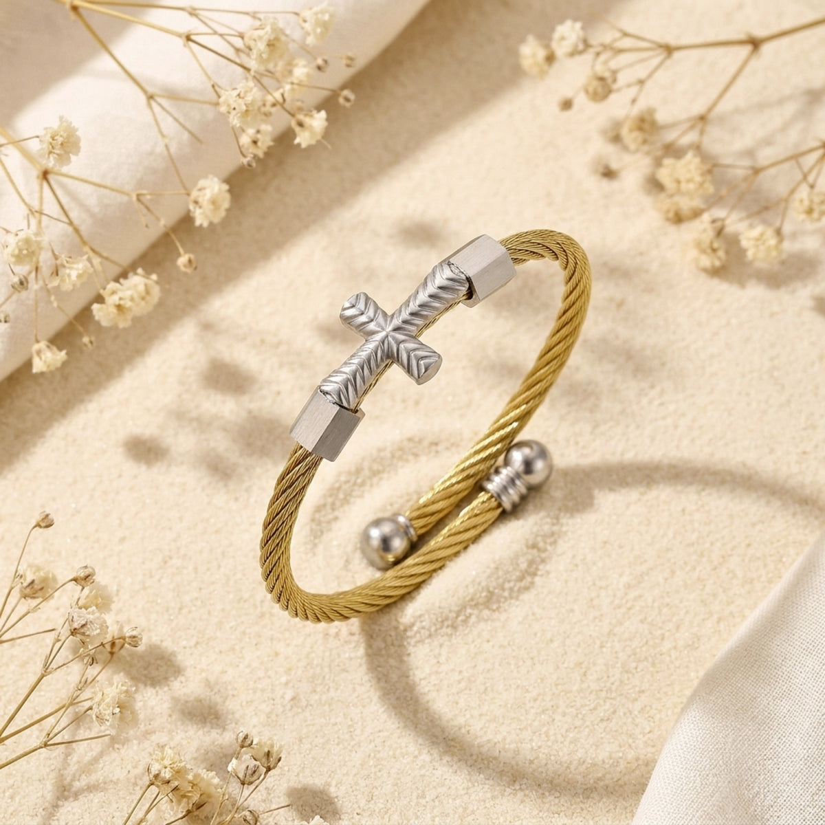 Gold-tone twisted cable cuff bracelet with a silver-tone cross accent on sand among dried flowers.
