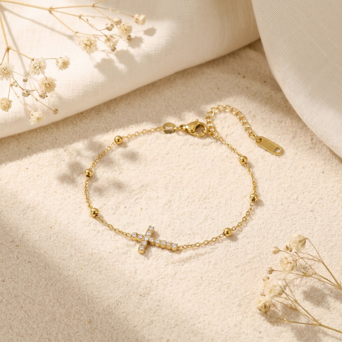 Gold-tone chain bracelet with a pave cross station laid on sand beside white fabric and dried flowers.