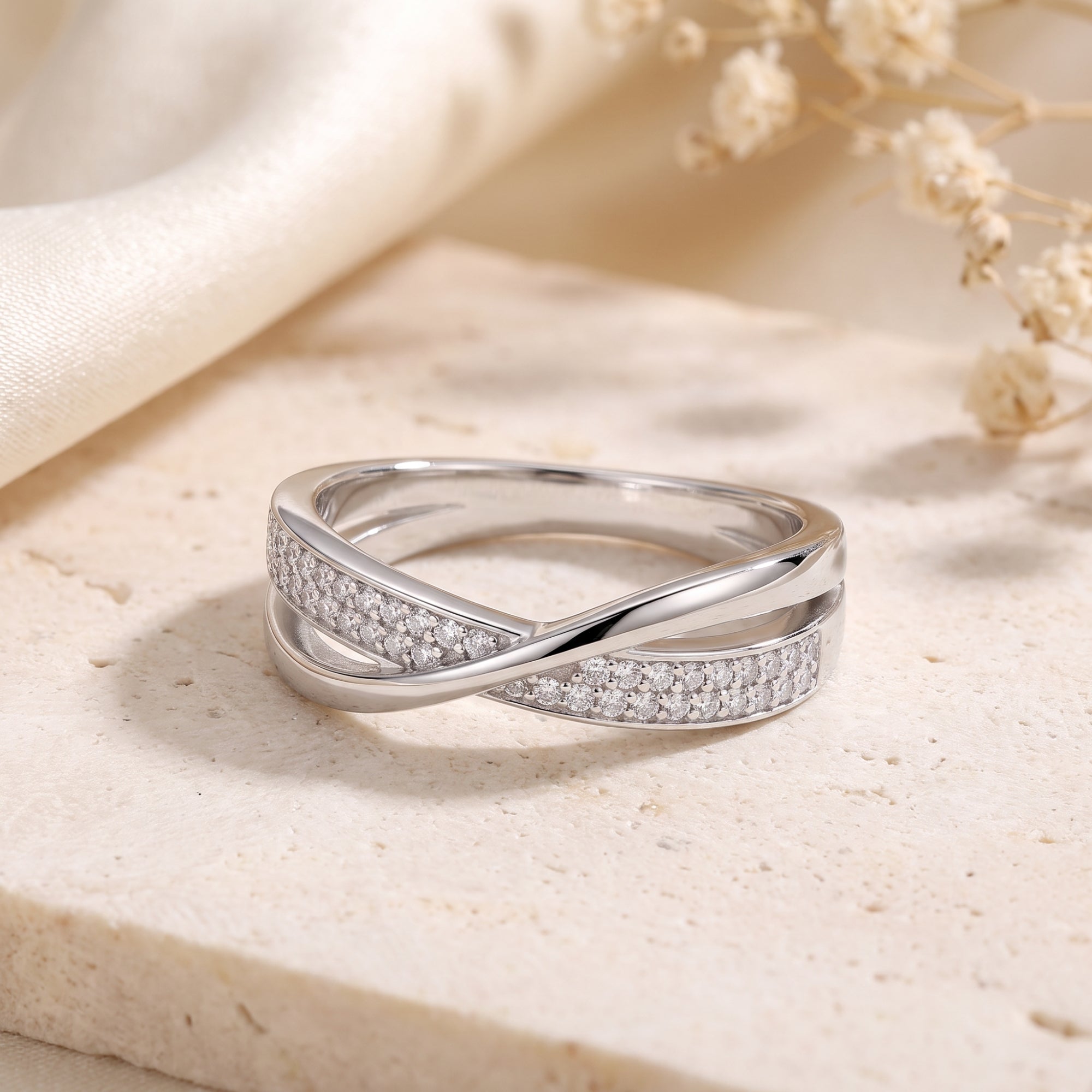 Silver crossover ring with pavé stones on a stone surface with dried flowers.