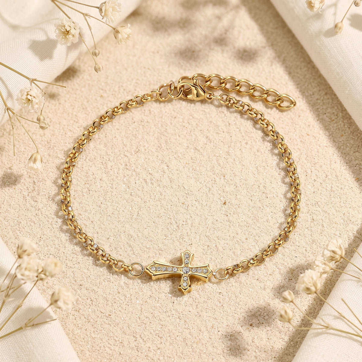 Top-down view of a gold-tone cross bracelet with sparkling stones on a sandy surface among white dried flowers.