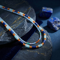Close-up of a layered solid yellow gold necklace with 5mm natural lapis lazuli beads on a dark stone background.
