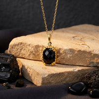 Yellow gold necklace with a cushion-cut black onyx pendant, styled on light-colored stones with raw minerals.