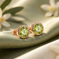 Round cut natural peridot stud earrings in 18K rose gold vermeil bezel settings on green silk with white flowers.