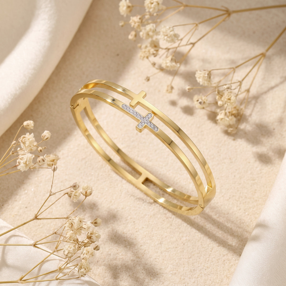 Gold-tone double-band cross bangle set diagonally on textured fabric and sand beside dried flowers.