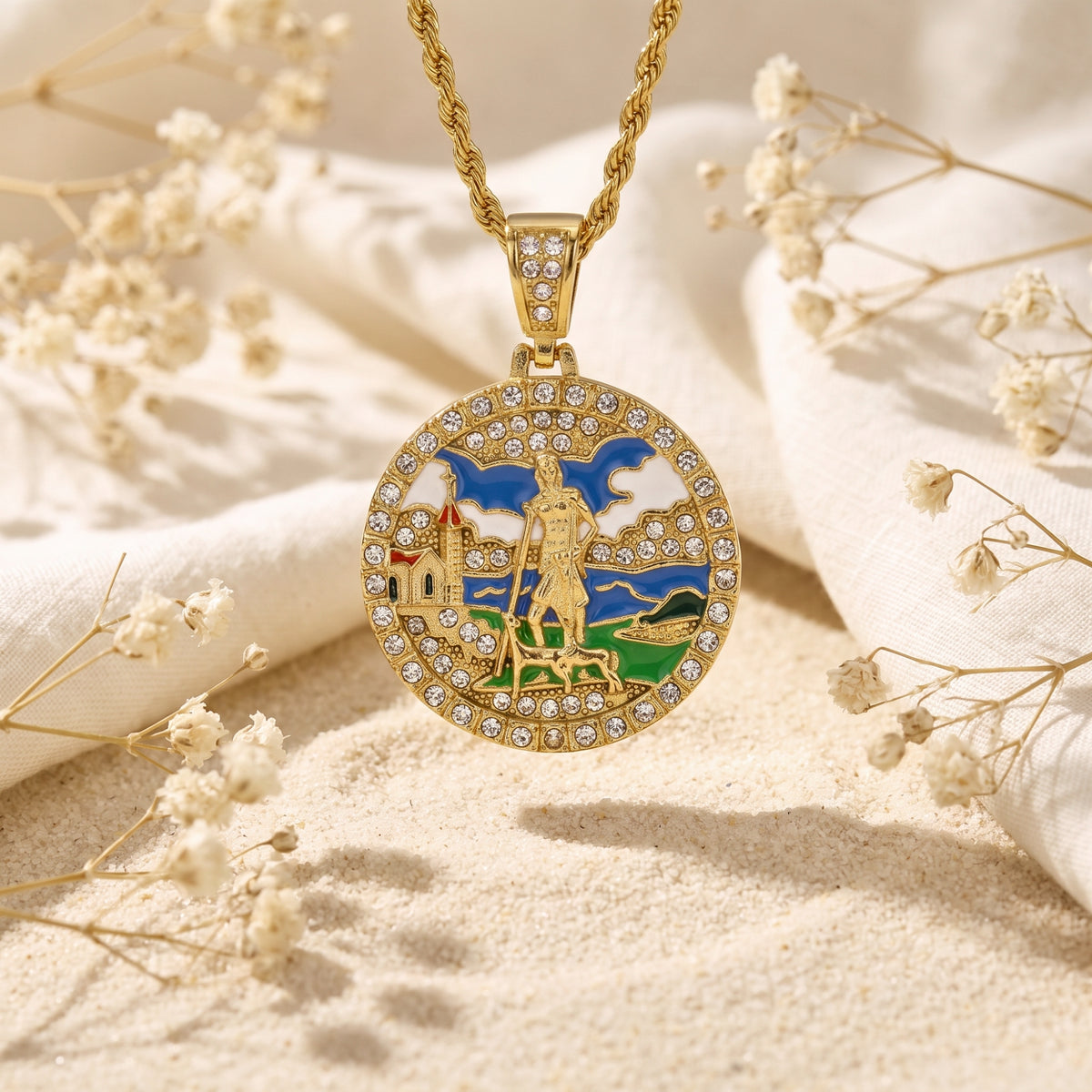 Gold-tone circular enamel medallion pendant with clear crystals, suspended over sand among dried flowers.