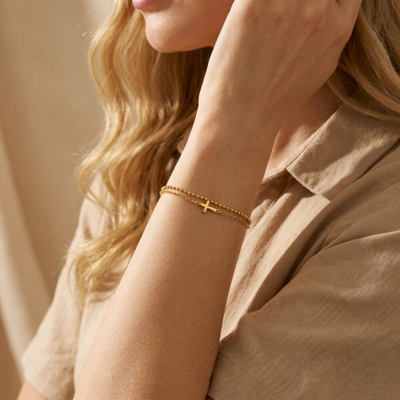 Gold-finish double-chain cross bracelet worn on a woman's wrist beside a beige collared shirt.