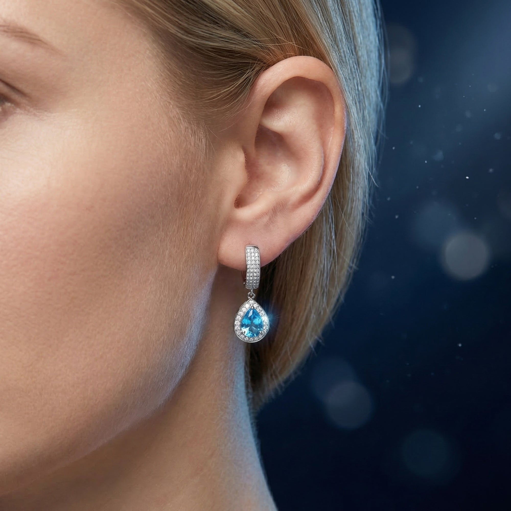 A pear cut Swiss blue topaz drop earring in a silver halo setting worn on a woman's ear.