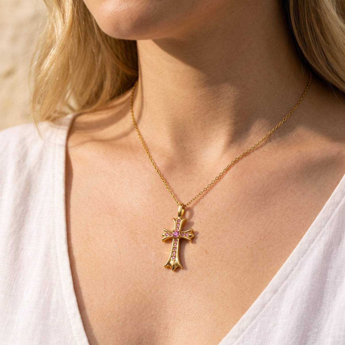 Gold-tone cross necklace with pink stones worn at a model's collarbone above a white top.