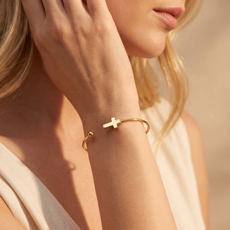 Close-up view of a gold-tone cross cuff bracelet worn on a woman's wrist against neutral clothing.