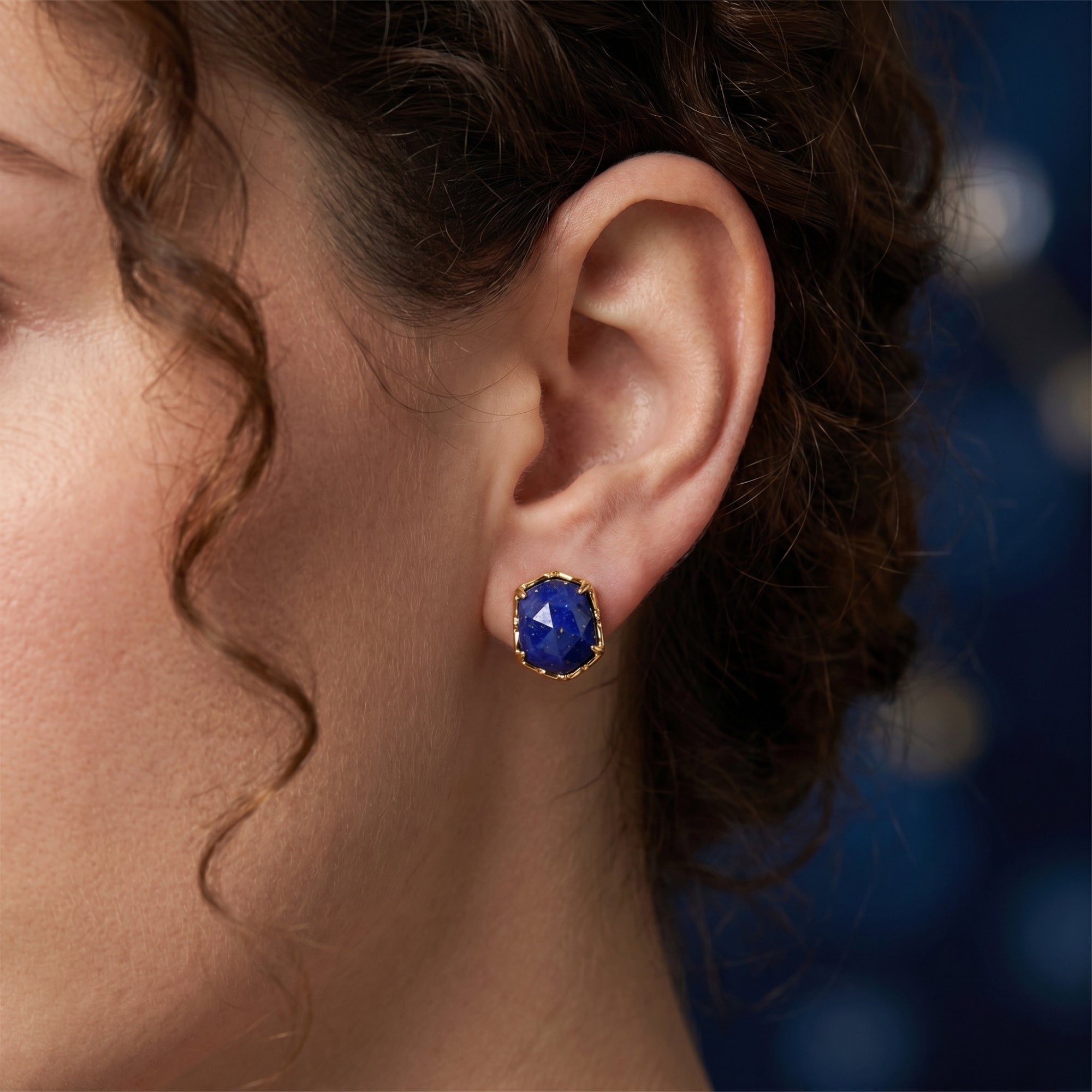 A woman wearing a gold hexagon stud earring featuring a natural blue lapis lazuli gemstone.