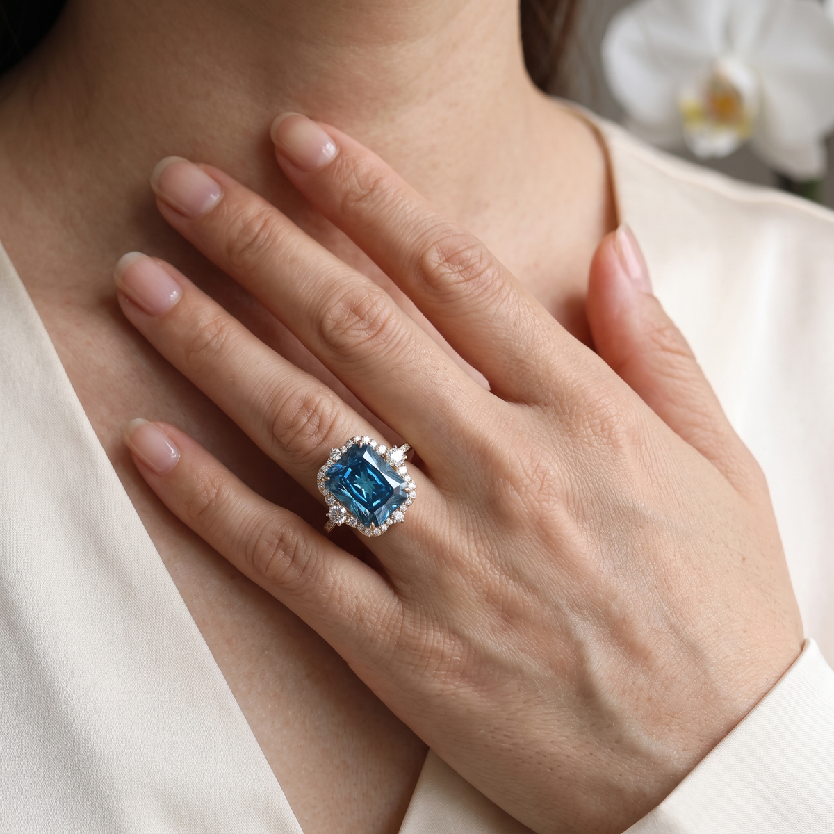 Close-up of a radiant cut blue moissanite halo ring in rose gold worn on a hand.