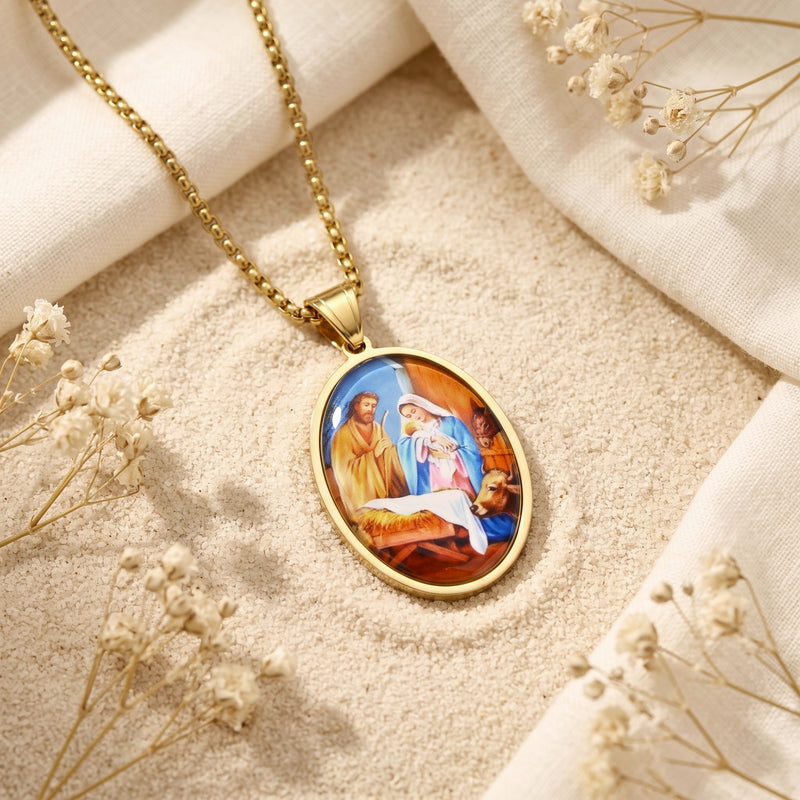 Gold-tone oval Nativity pendant necklace lying diagonally on pale sand beside white fabric and dried flowers.