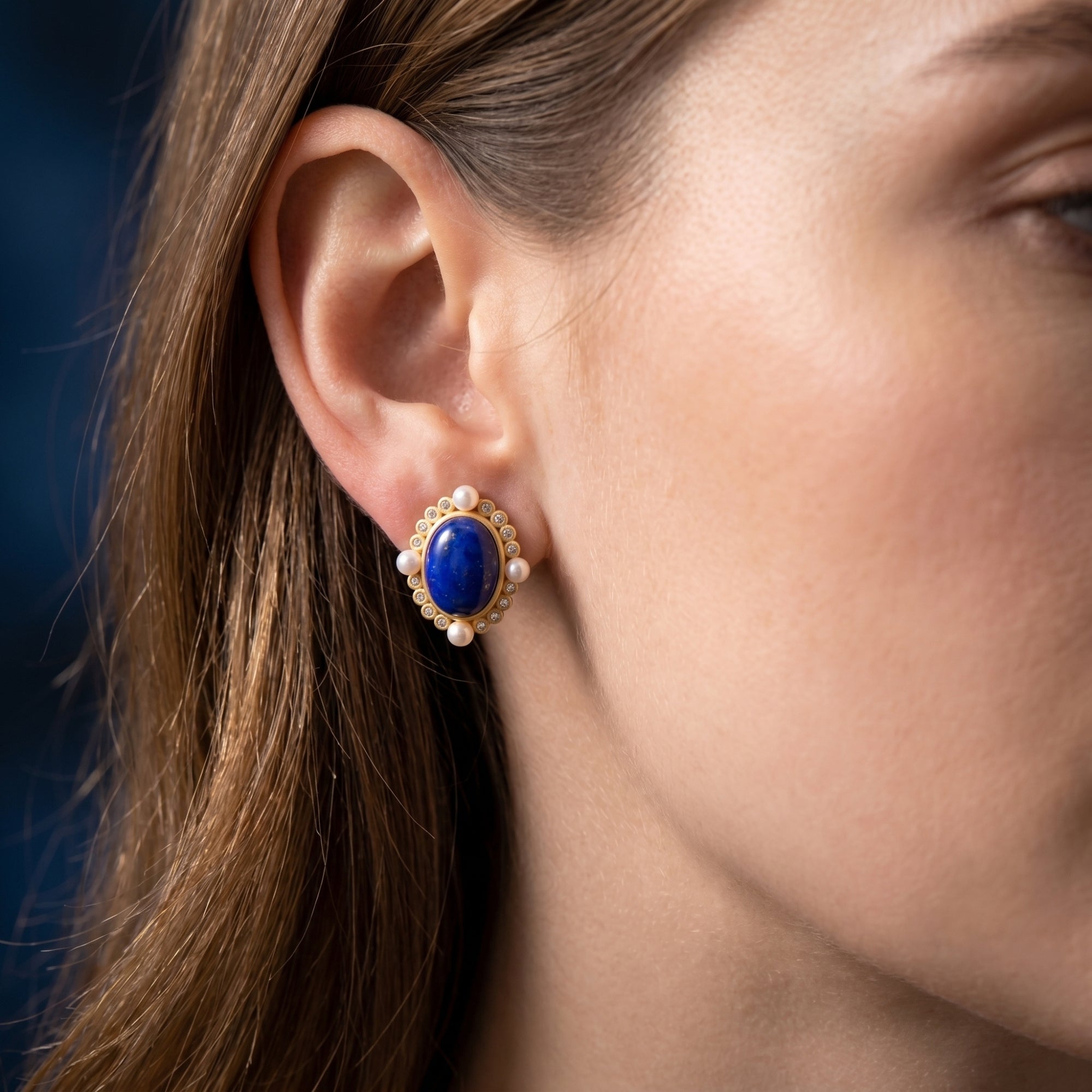 Close-up of a woman's ear wearing a gold-plated oval lapis lazuli stud earring with pearls and crystals.