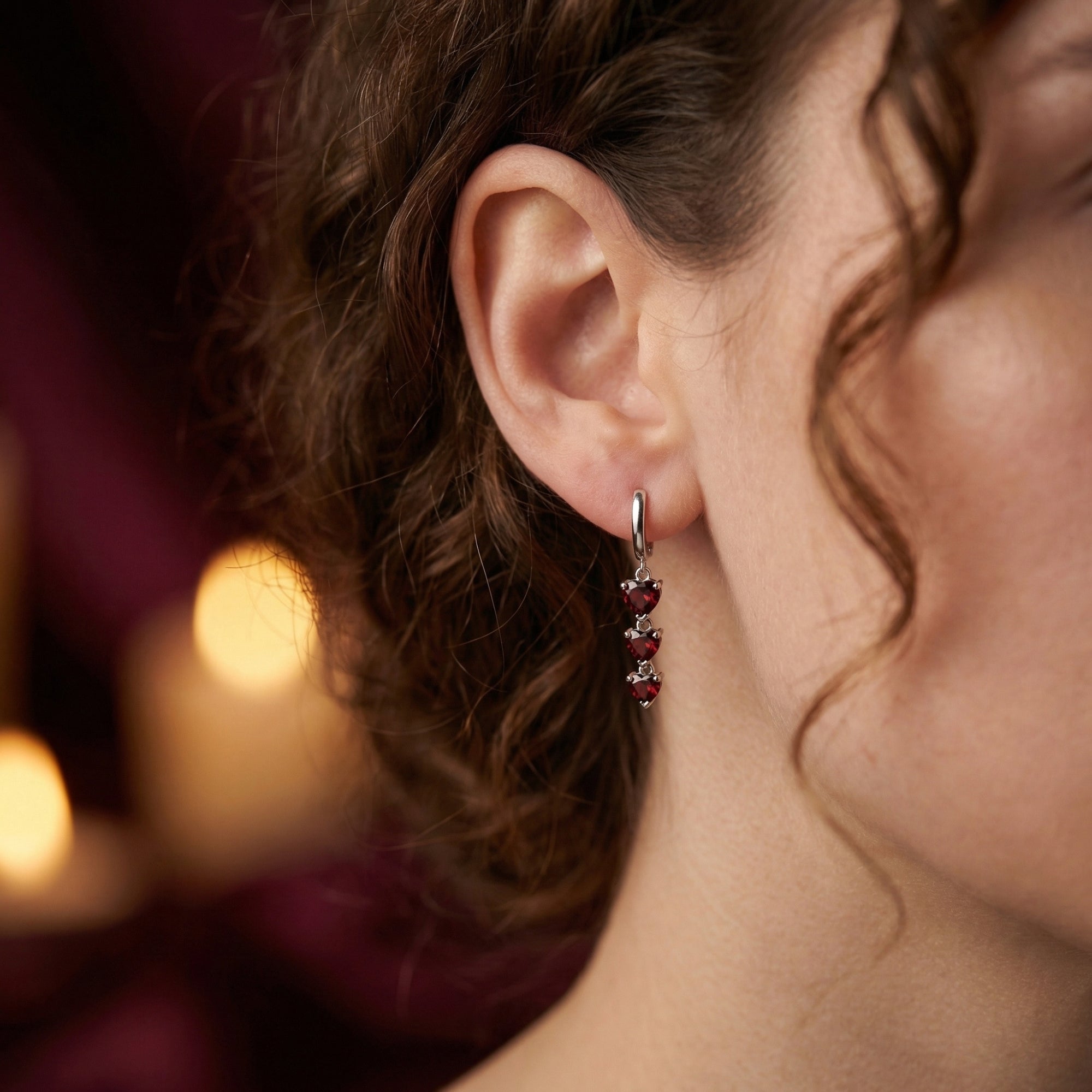 Model wearing a silver heart drop earring with three cascading heart-cut red garnets against a soft-lit background.
