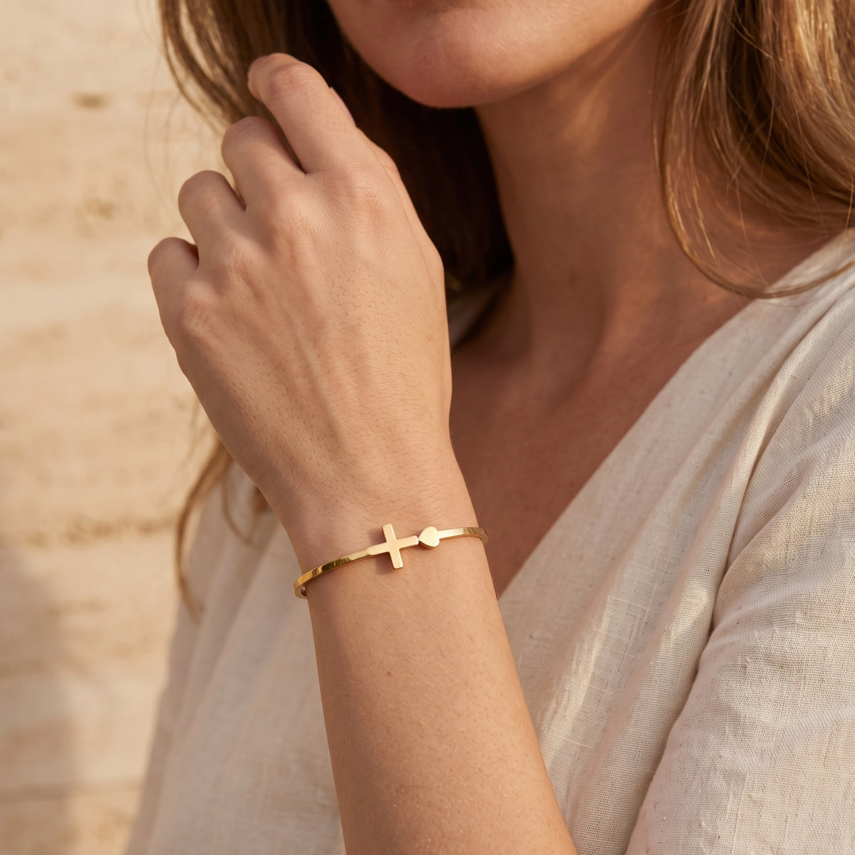 Gold-tone cross and heart bangle bracelet worn on a woman's wrist with her hand raised near her face.