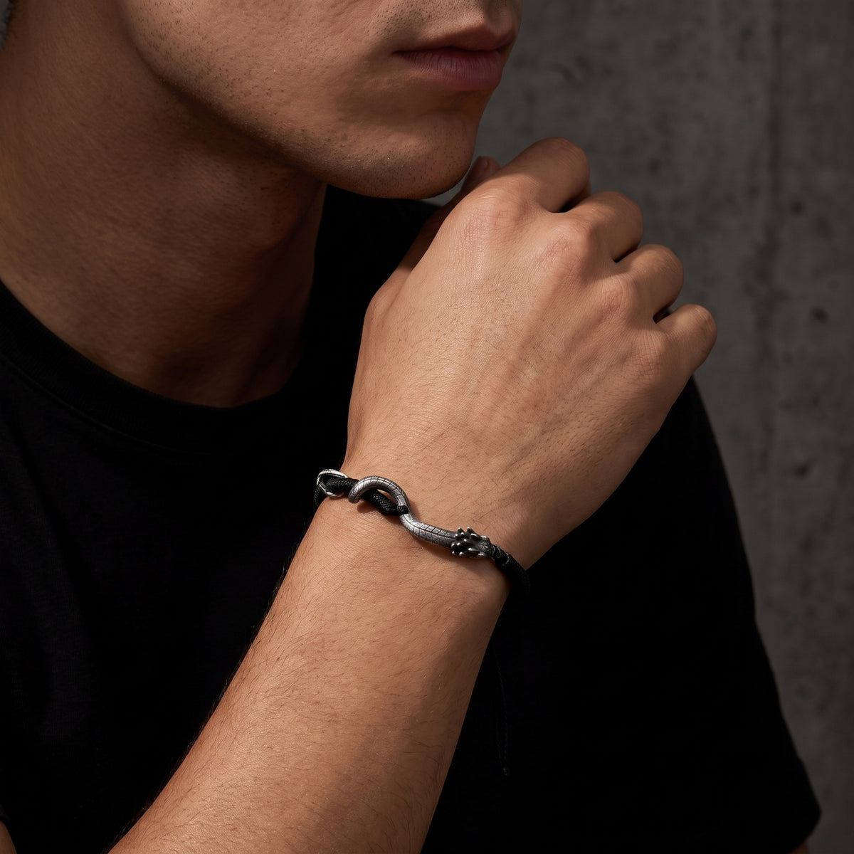 Man wearing a black cord bracelet with a silver serpent centerpiece, his wrist raised near his chin.