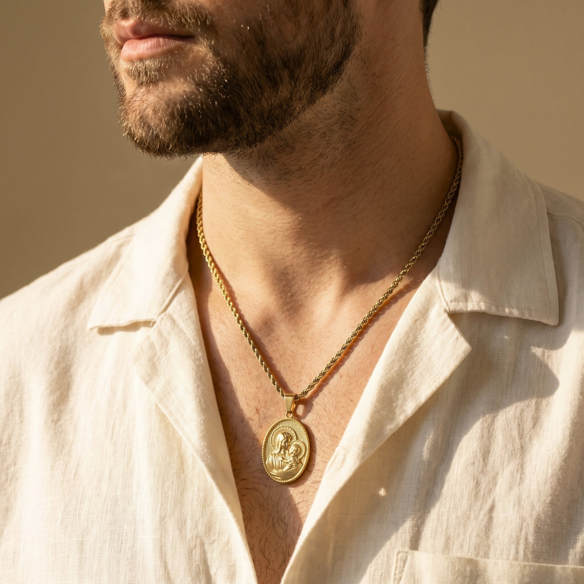 Gold-tone Madonna medallion necklace worn on a man in a cream linen shirt.