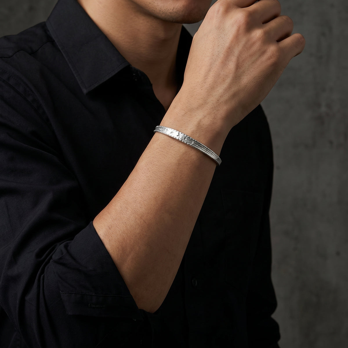 Etched silver cuff bracelet worn on a raised wrist over a black button-down shirt.