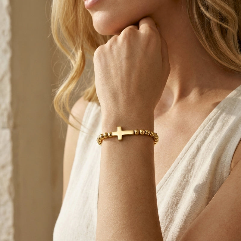 Model wearing a gold-tone beaded cross bracelet on her wrist with her hand resting beneath her chin.