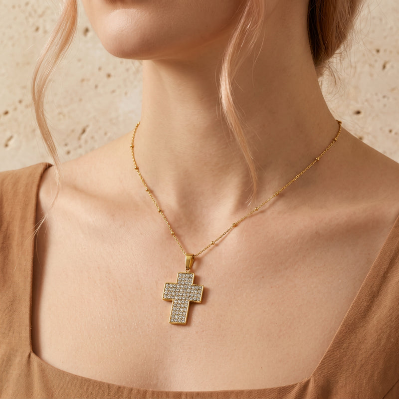 Model wearing a gold-tone pave cross pendant necklace over a brown square-neck top.