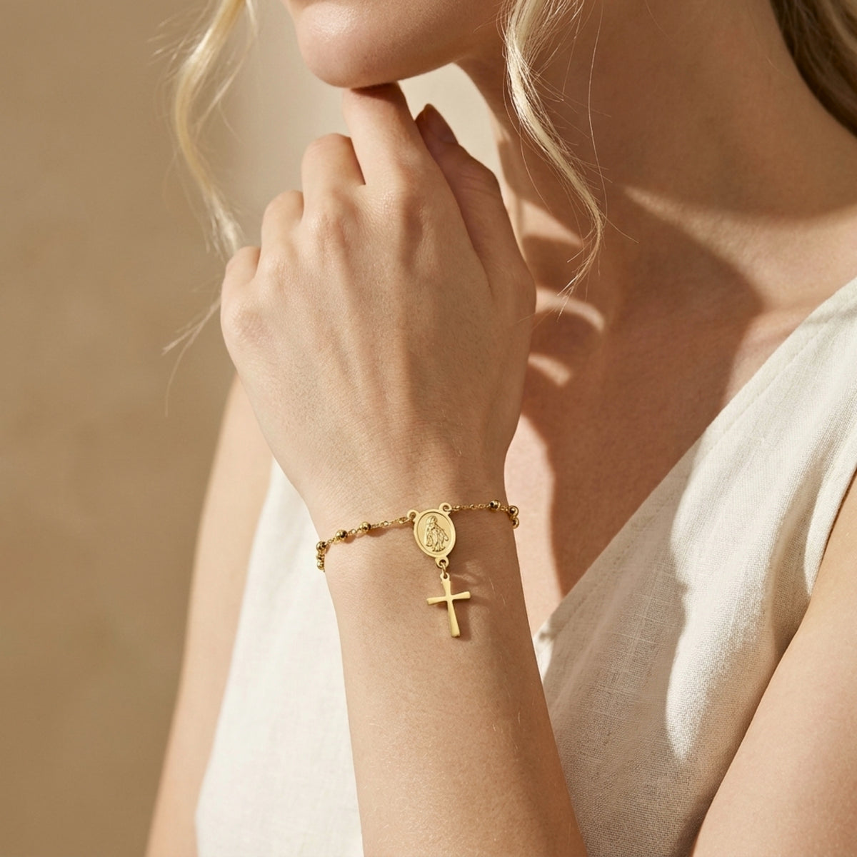 Gold-tone rosary bracelet worn on a wrist, with a Virgin Mary medal and dangling cross charm; hand rests near the face.