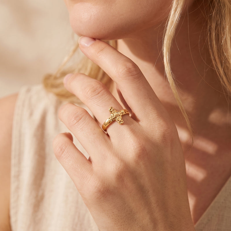 Gold-tone sculpted cross ring worn on a woman's finger in a close-up near her face.