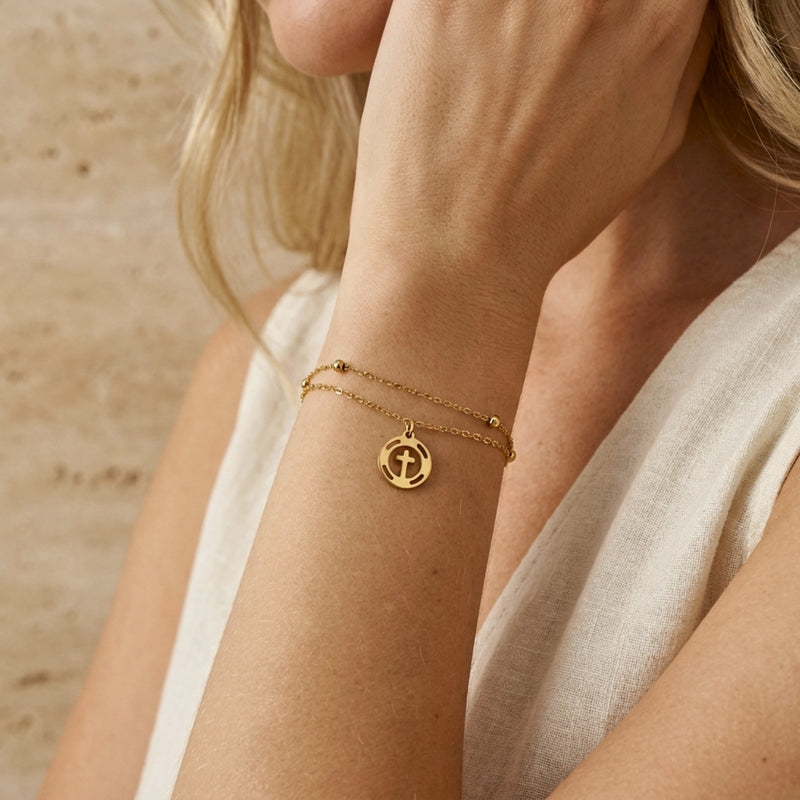 Close-up of the double-strand gold-tone cross bracelet worn on a woman's wrist over white clothing.