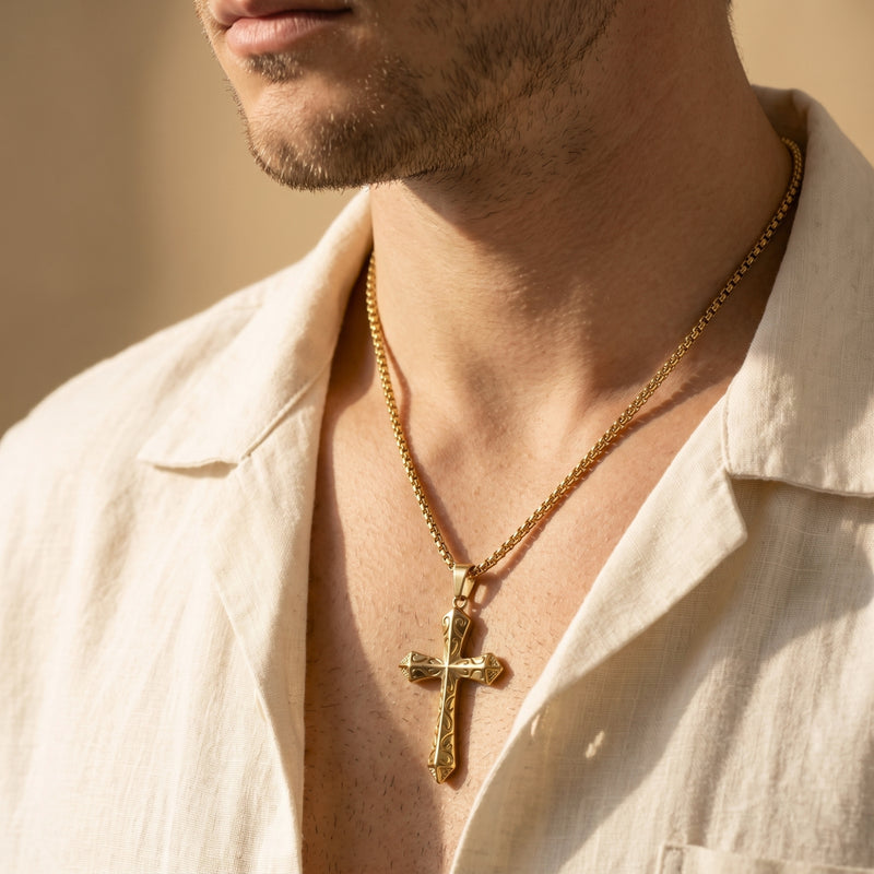 Close-up of a man wearing the gold-tone scrollwork cross pendant on a box chain over an open linen shirt.