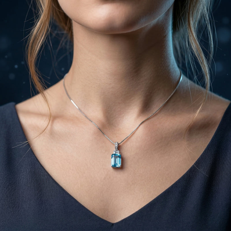 Model wearing the Coraline emerald cut blue topaz necklace in silver against a dark blue background.
