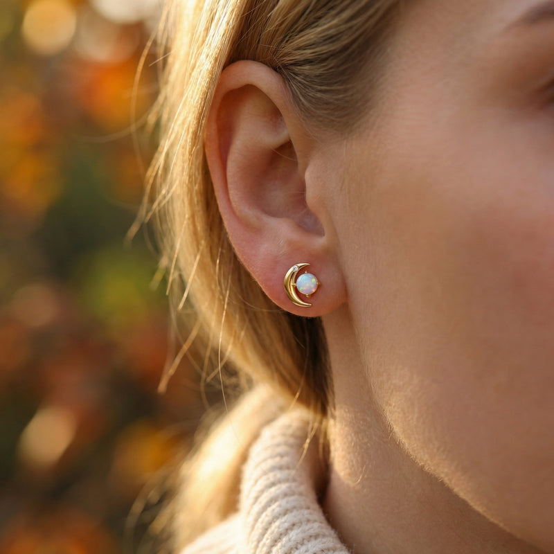 Woman wearing a gold-tone crescent moon stud earring with a round iridescent opal stone in an outdoor setting.
