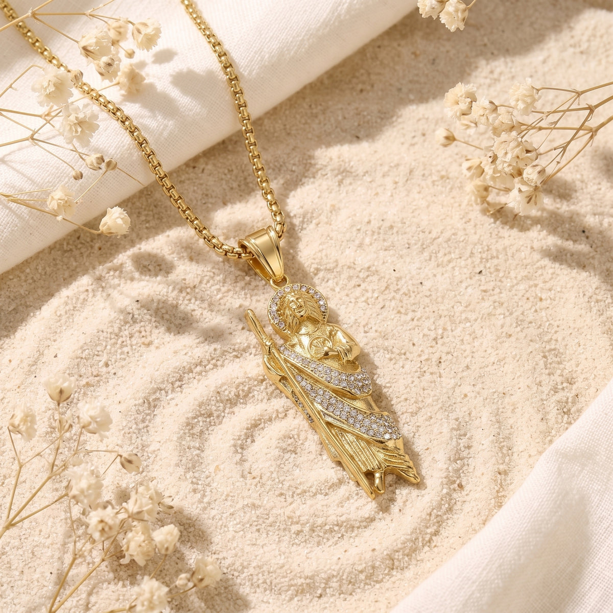 Gold-tone St. Jude pendant necklace lying diagonally on sand among dried white flowers.