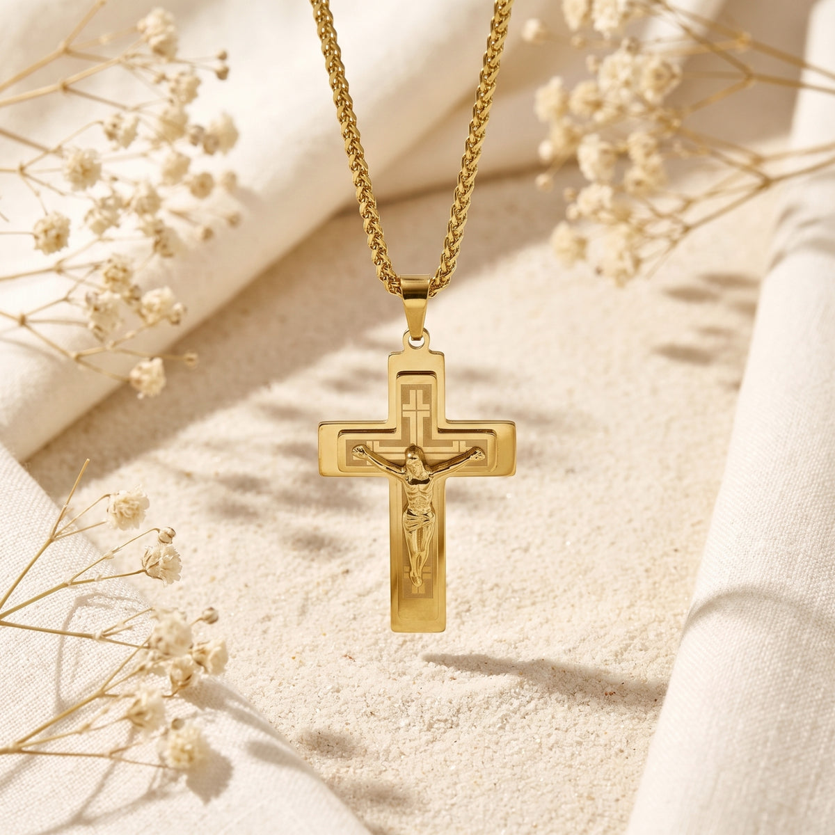 Gold-tone layered crucifix pendant hanging vertically from its chain over sand and white fabric
