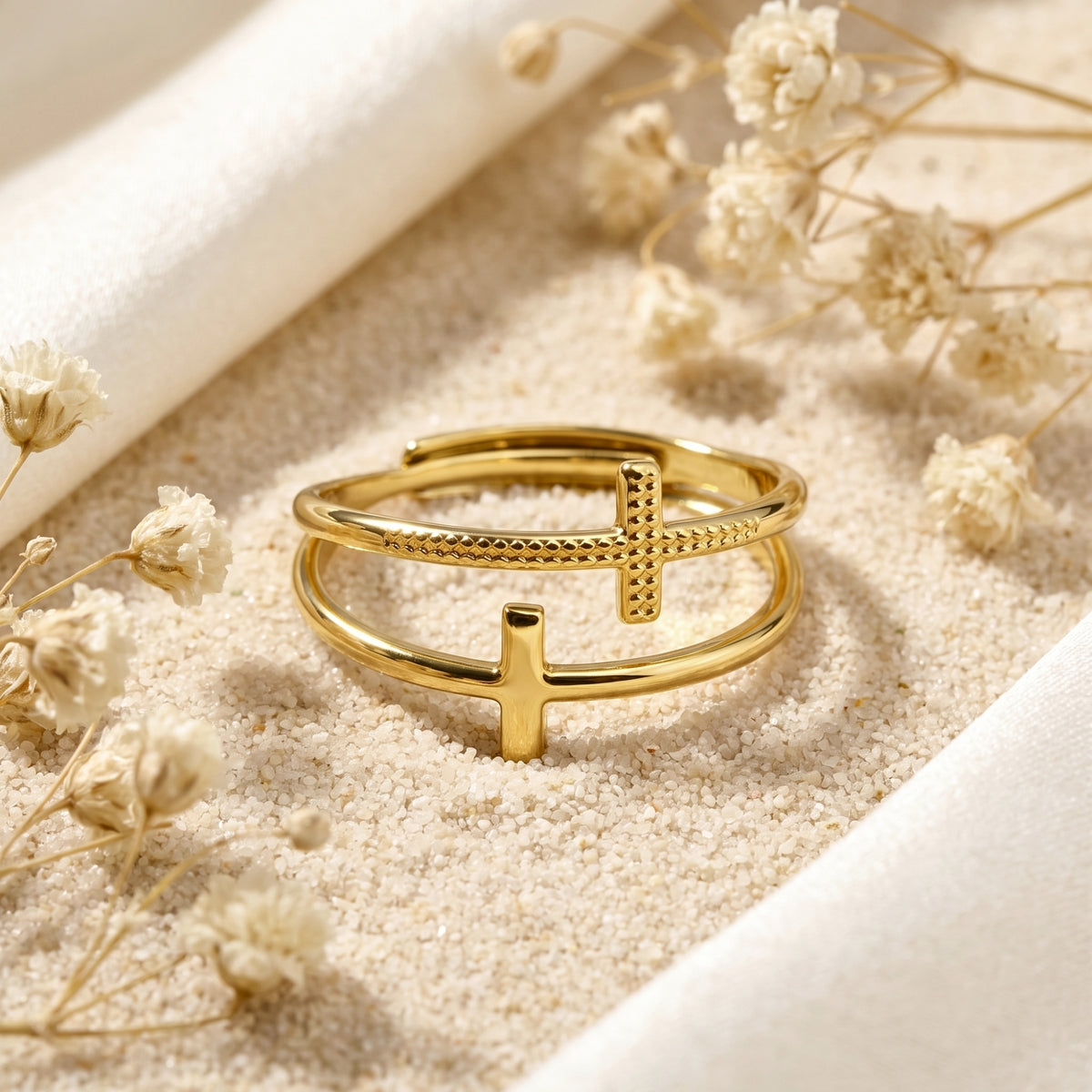 Gold-tone double cross ring displayed horizontally on sand with white dried flowers.