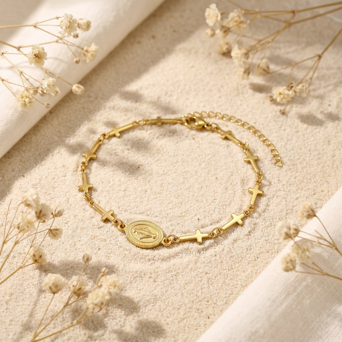 Angled view of a gold-tone cross station bracelet with an oval medallion displayed on sand.