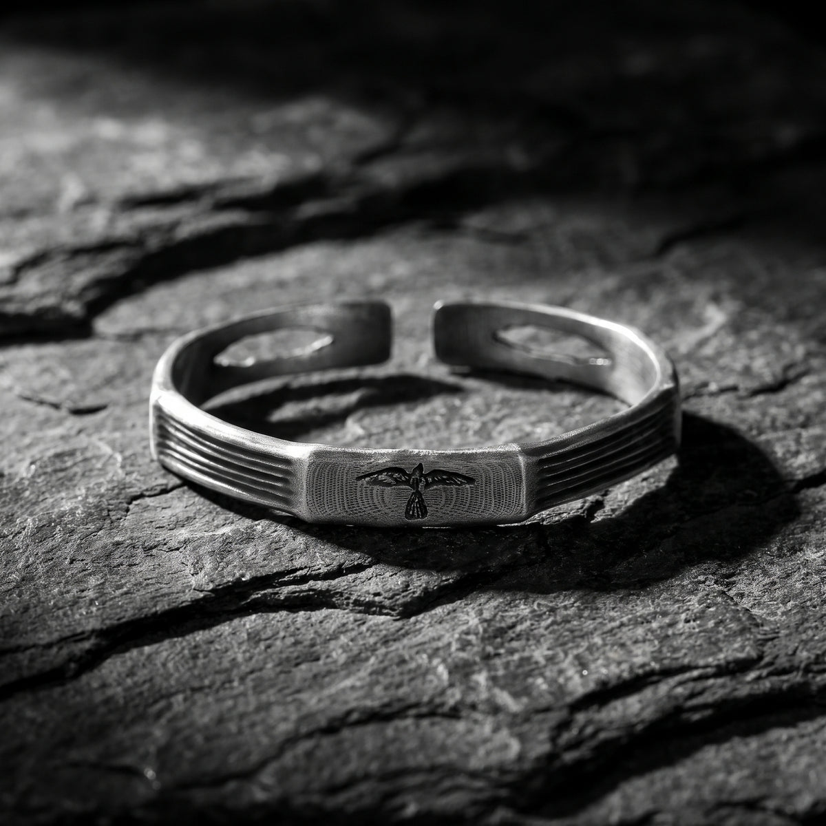 Front view of the open silver cuff bracelet on dark stone, showing the etched raven plaque.