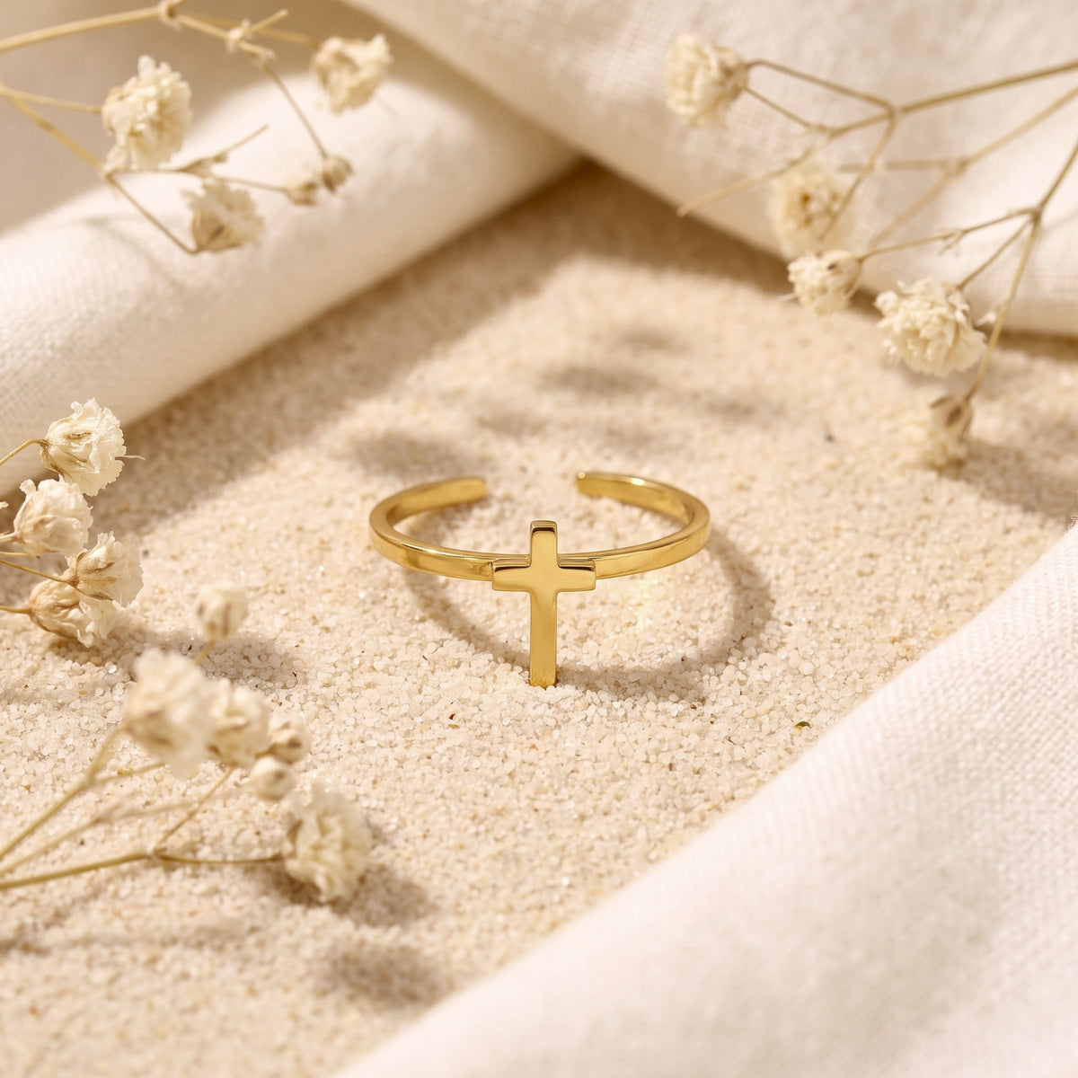 Front view of the gold-tone open cross ring standing in sand among white fabric and dried flowers.