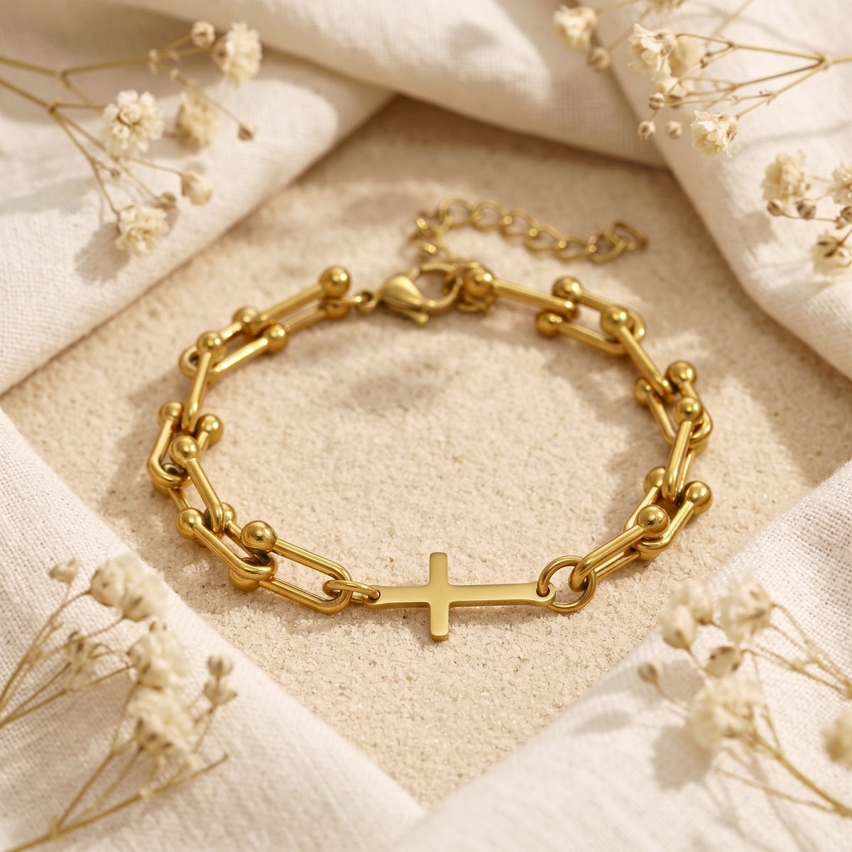 Gold-tone U-link bracelet with a horizontal cross displayed on sand, framed by draped white fabric.