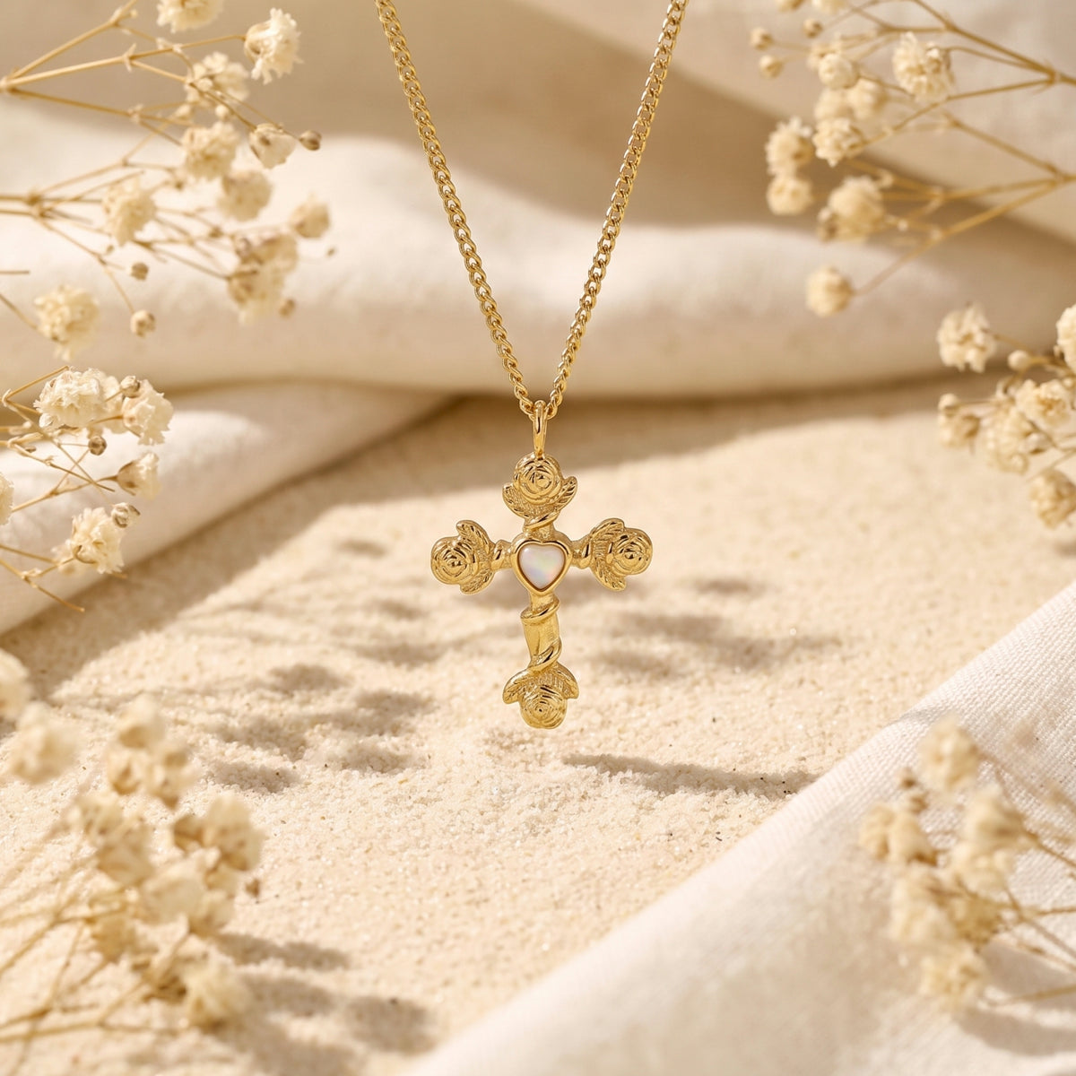 Gold-tone rose cross necklace hanging vertically over sand with dried flowers around it.