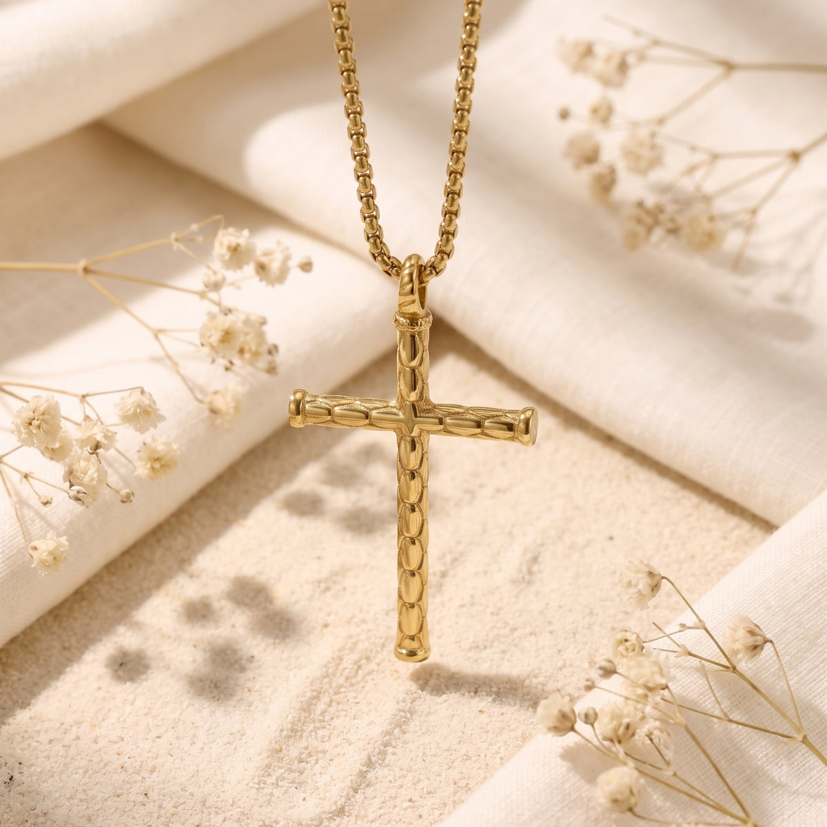 Gold-tone segmented cross necklace suspended above sand and white fabric with dried flowers.