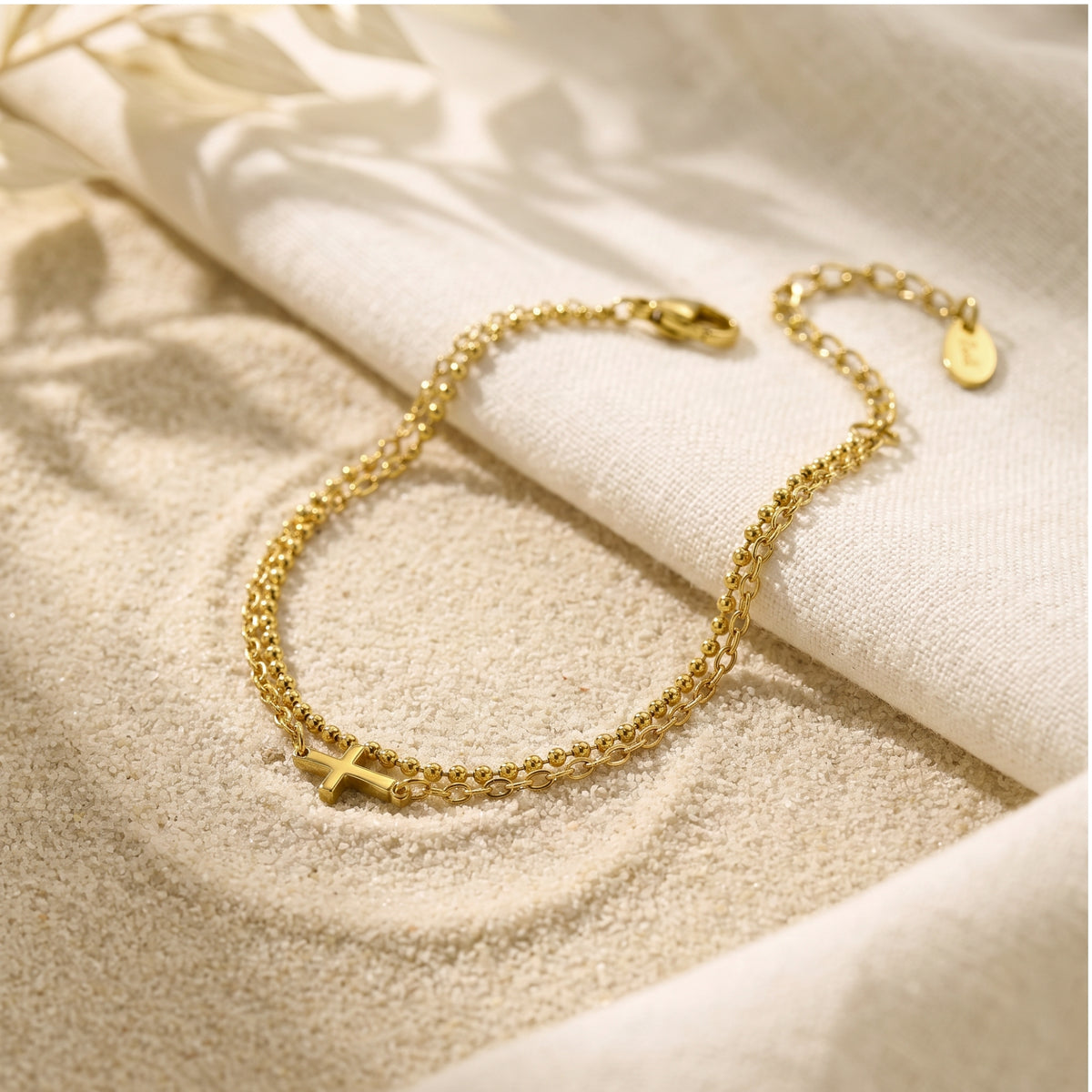 Angled view of a gold-finish double-chain cross bracelet on sand beside white linen.